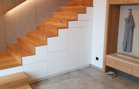 Das Bild zeigt eine Treppe aus Buchenholz, die zu einem höheren Bereich führt. Unter der Treppe sind weiße Schränke eingepasst, die zusätzlichen Stauraum bieten. Auf der rechten Seite ist eine Garderobe zu sehen.