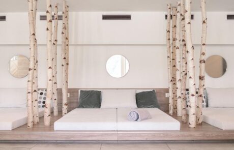 Auf dem Bild ist ein Wellnessbereich zu sehen, in dem Doppelbetten nebeneinander platziert sind. Die Betten werden durch kunstvolle Baumstämme, die vom Boden bis zur Decke reichen, voneinander getrennt. Der Raum ist sehr hell gestaltet und lässt viel natürliches Licht herein. Die Betten befinden sich auf einem Podest, das aus Eichenholz gefertigt ist. Das Holz verleiht dem Raum eine natürliche und warme Atmosphäre. Der Stil des Raumes ist schlicht und elegant, ohne ausschmückende Details. Es geht um eine klare, minimalistische Ästhetik, die zur Entspannung und Erholung einlädt.