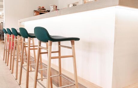 Theke im Hotel "Villa Postillon". Die Theke ist in Weiß gehalten und bildet einen Kontrast zu dem Sockel und der Tischplatte, die aus Eiche gefertigt sind. Direkt vor der Theke stehen fünf Hocker